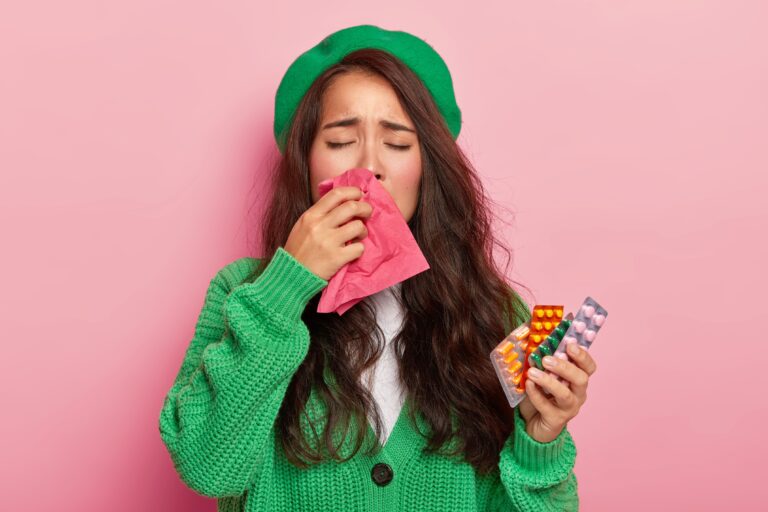 Unhappy brunette girl suffers from flu symptoms, rubs nose with handkerchief, has cold, holds pills, wears green jumper and cap, isolated on pink wall.