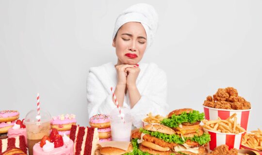 Binge eating concept. Stressed unhappy Asian lady wants to eat fast food tempting and looks with appetite has addiction to cheat meal wears comfortable bathrobe towel over head poses indoor.