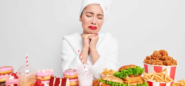 Binge eating concept. Stressed unhappy Asian lady wants to eat fast food tempting and looks with appetite has addiction to cheat meal wears comfortable bathrobe towel over head poses indoor.